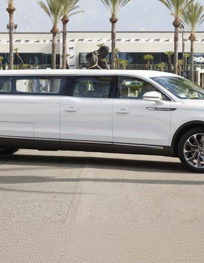 Side profile of a white 6-door GMC Yukon Denali SUV parked in front of palm trees, featuring extended wheelbase and chrome accents.