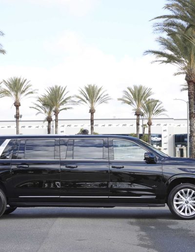 Full side view of a black 6-door GMC Yukon Denali SUV conversion, parked in front of palm trees at Quality Coachworks.