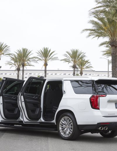 Rear three-quarter view of a white 6-door GMC Yukon Denali SUV with all passenger doors open, showcasing extended access.