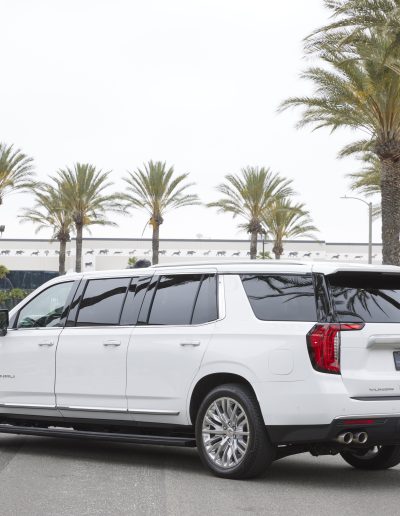 Angled rear view of a white 6-door GMC Yukon Denali conversion parked outside Quality Coachworks, featuring full-length body design.