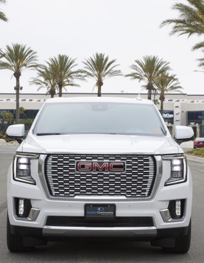 Frontal view of a white 6-door GMC Yukon Denali SUV parked in front of palm trees, converted by Quality Coachworks for VIP or group transport.