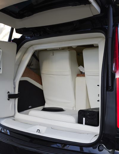 Open rear hatch of a Cadillac Escalade VIP conversion showing white paneling and concealed storage compartment by Quality Coachworks.