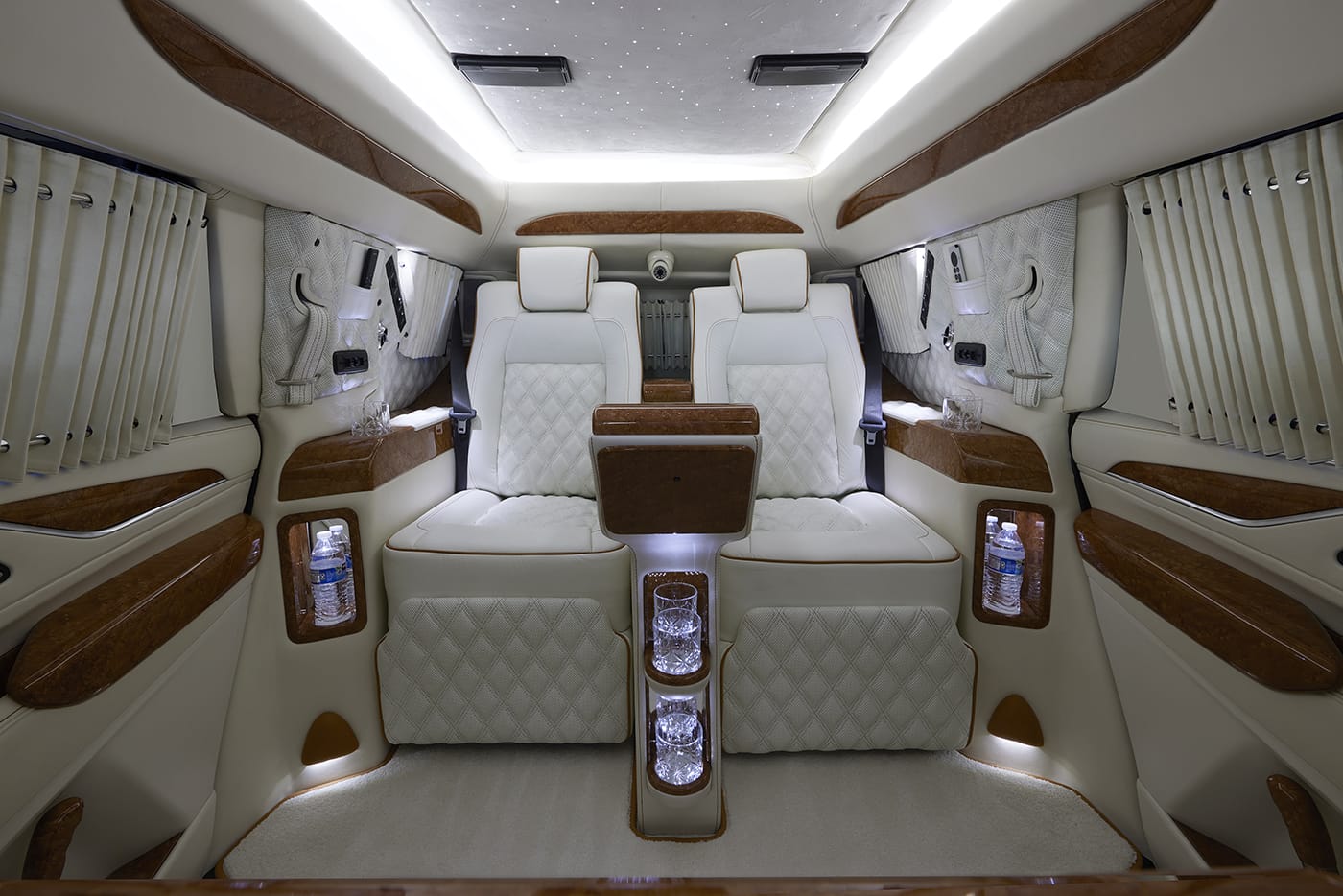 Interior of a luxury armored vehicle featuring two plush white diamond-stitched captain chairs with tan leather trim, a wood-accented center console with built-in glass holders, LED accent lighting, privacy curtains on the windows, and recessed compartments holding bottled water. The ceiling includes a starlight headliner, creating an upscale, executive atmosphere.