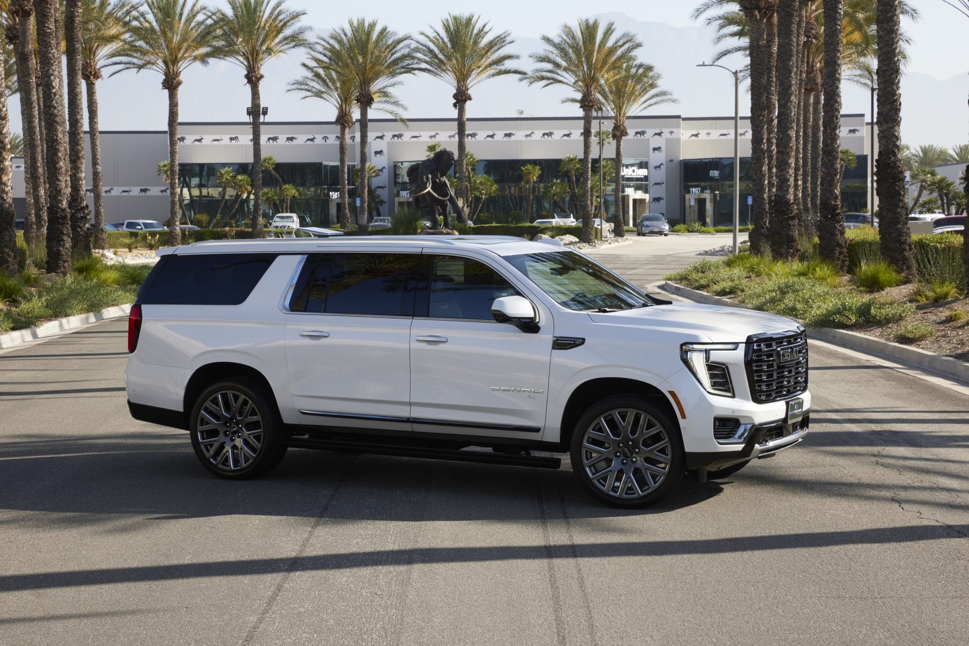 A 2025 white GMC Yukon Denali Ultimate SUV seen from a front‑three‑quarter view. It features a bold “Vader Chrome” grille exclusive to the Ultimate trim, distinctive C‑shaped LED headlights, body‑colored door handles, dual‑tone black/white side mirrors, and large premium wheels—visually identified as either 22″ or 24″ Ultra‑Bright machined aluminum rims in Black or After‑Midnight finish. The White Frost Tricoat paint emphasizes its premium stance, perched on an adaptive Air Ride suspension with the Denali badge visible on the front doors.